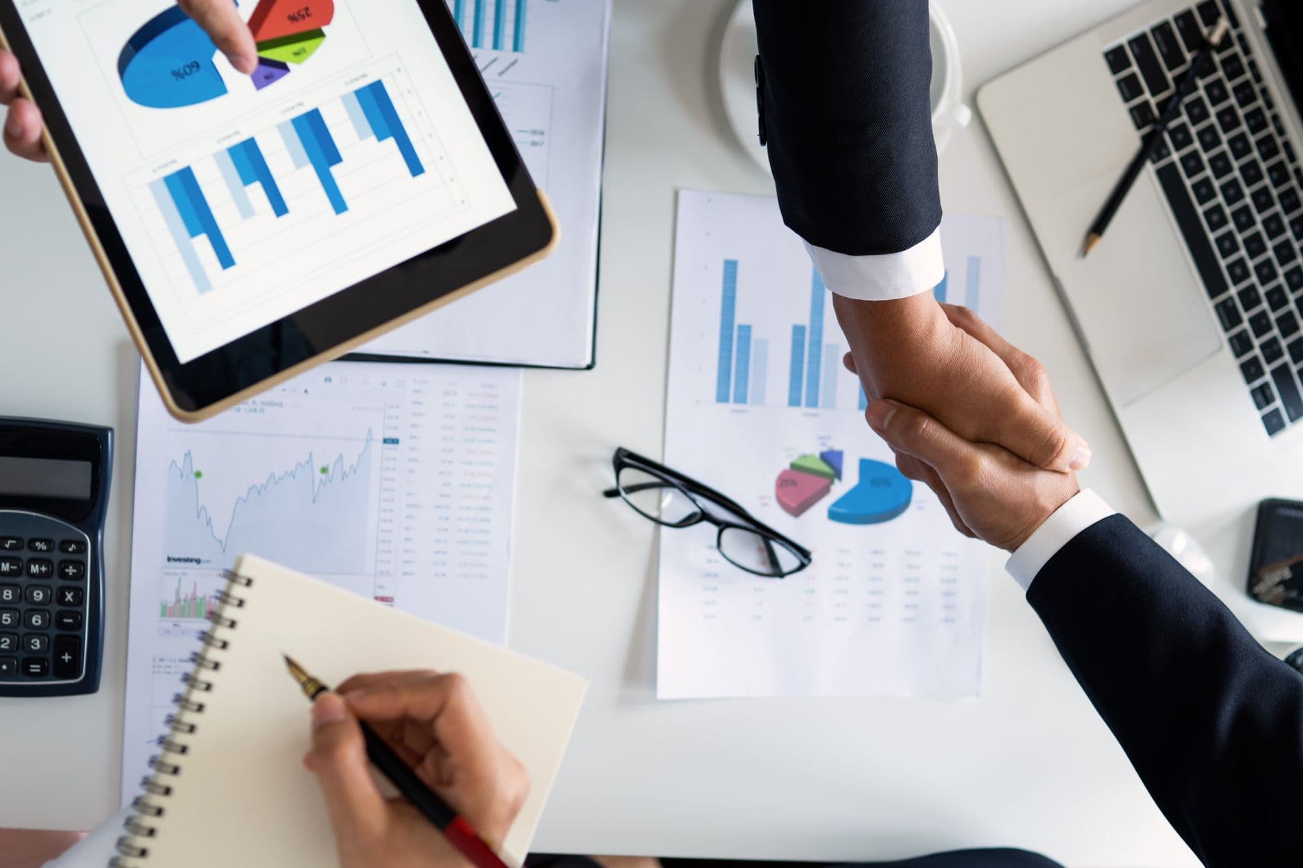 Two people shaking hands over a desk covered with printed charts, a tablet and a notepad